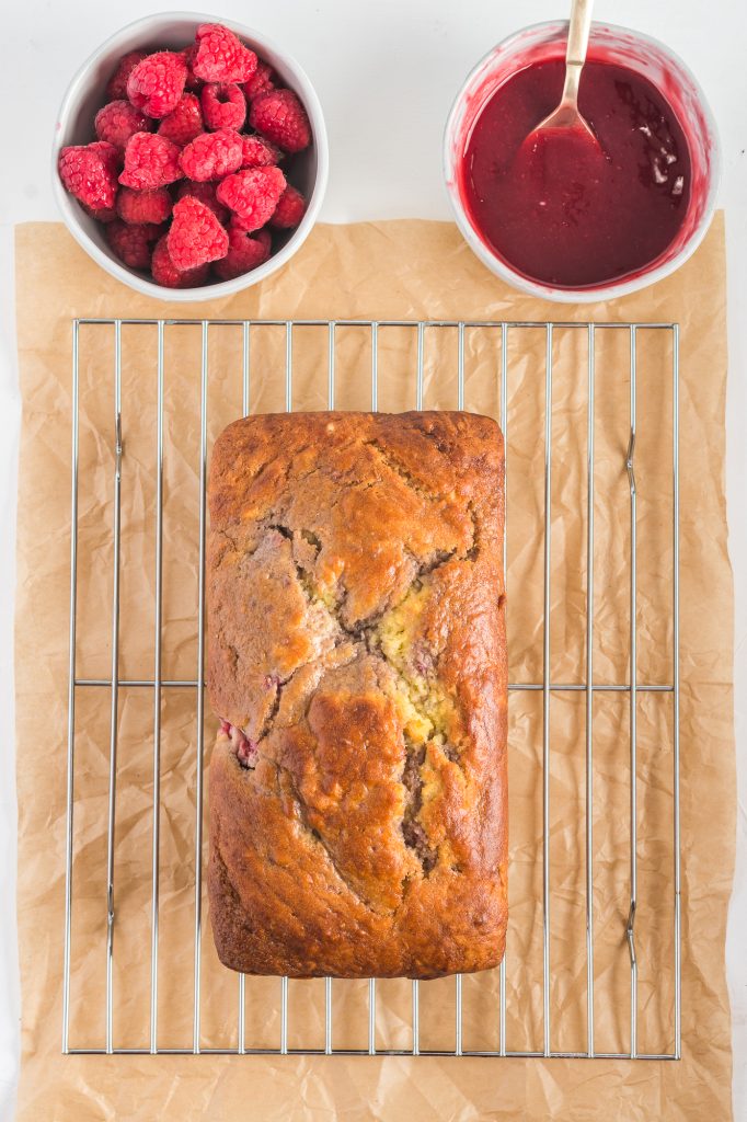 A freshly baked raspberry marble loaf sits on a cooling rack next to a bowl of raspberries and a bowl of raspberry glaze.