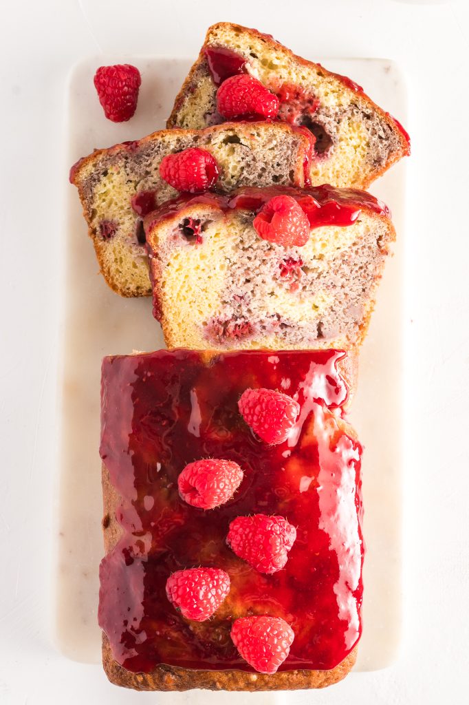 A raspberry marble loaf partially slice sits on a board and is decorated with a glaze and fresh raspberries.