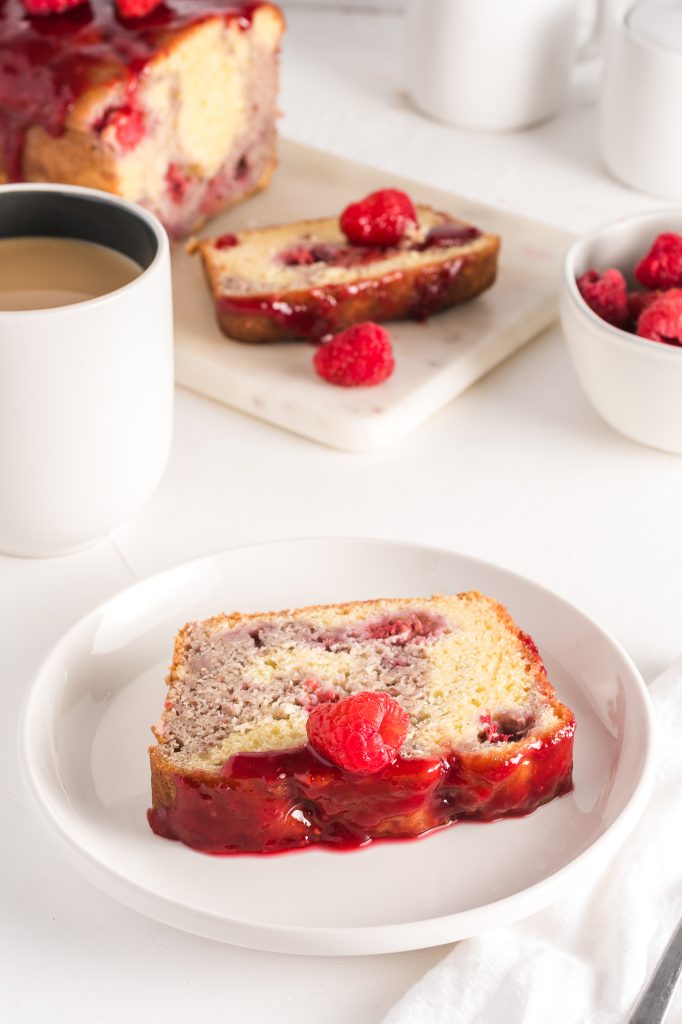 A single slice of raspberry marble loaf sits on a plate in the foreground with a cup of coffee, a bowl of raspberries, and the remainder of the loaf are in the background.