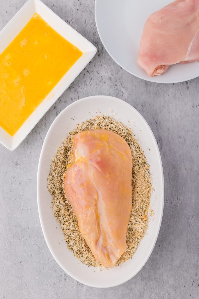 A slice of raw chicken coated in an egg mixture laying in a shallow bowl of spiced breadcrumbs.