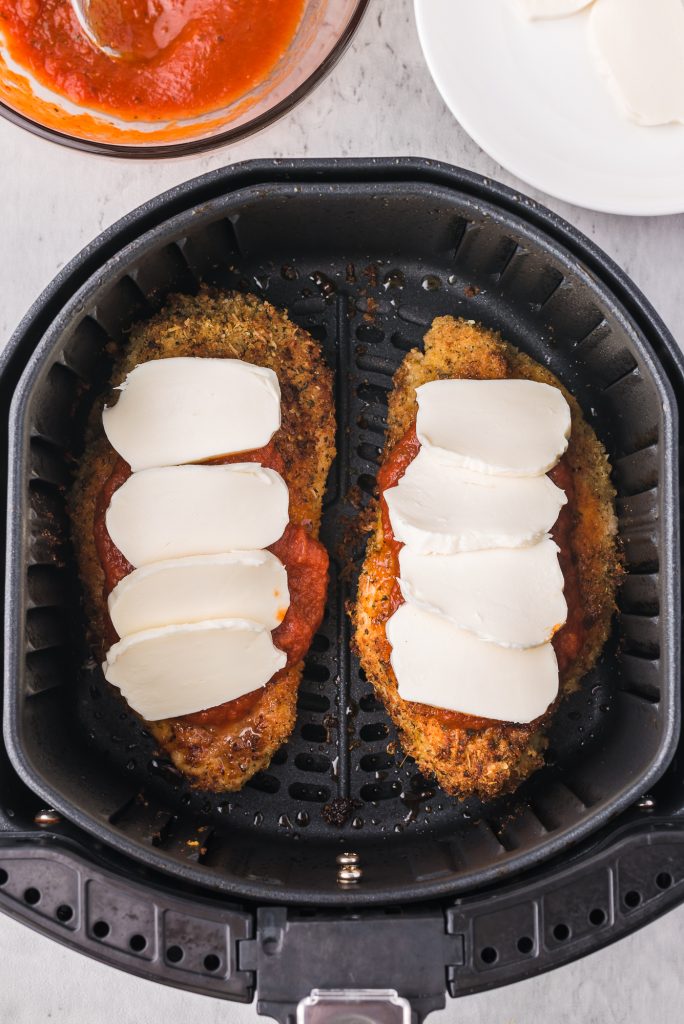 Two pieces of cooked, breaded chicken in an air fryer basket topped with marinara sauce and sliced mozzarella.