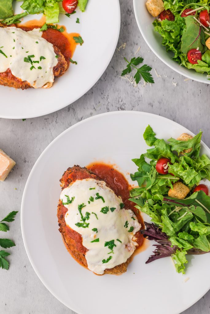 A white plate holds one piece of chicken parmesan and a green side salad. Two more plate are in the background.