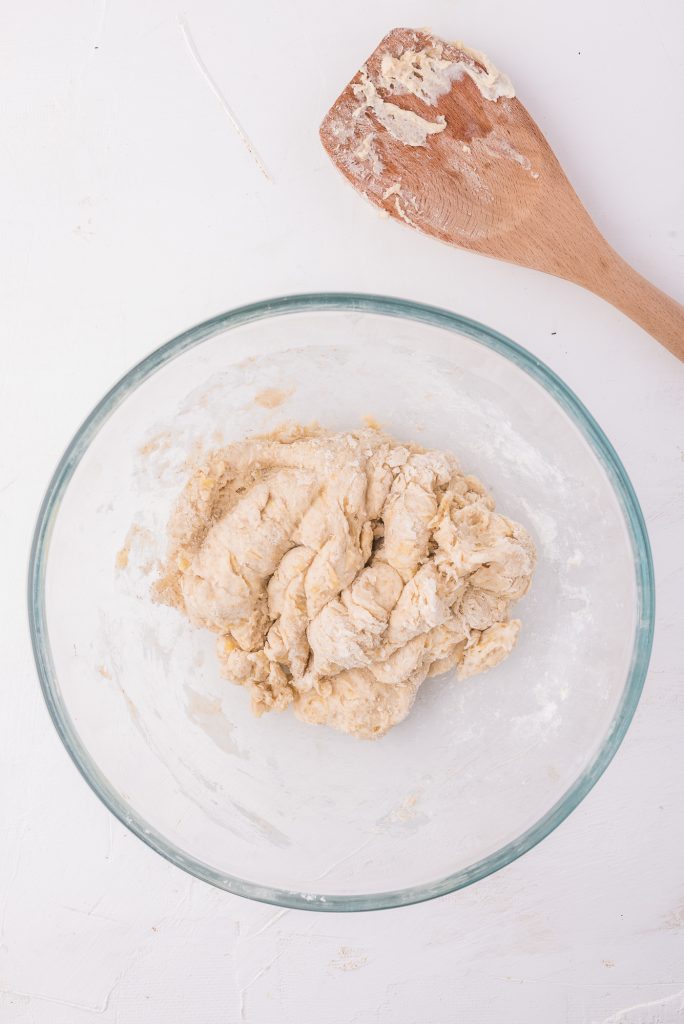 A glass bowl with a mixed dough and a wooden spoon next to it.