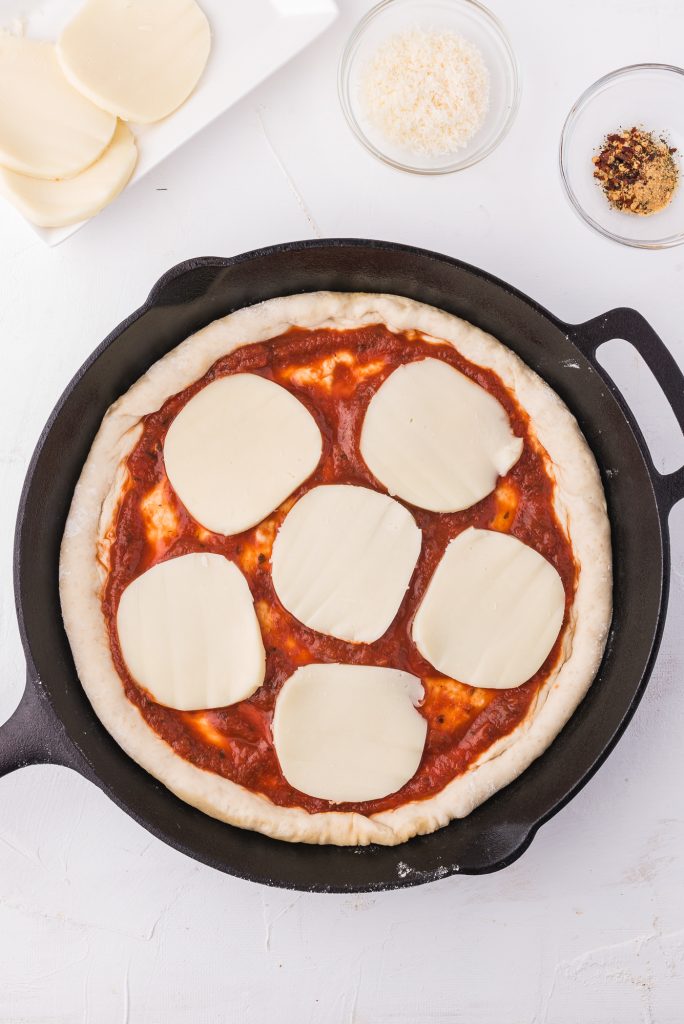 The pizza crust topped with pizza sauce and sliced mozzarella.