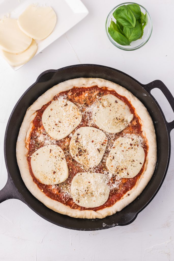 The pizza crust topped with pizza sauce, sliced mozzarella, and various spices from the ingredient list.