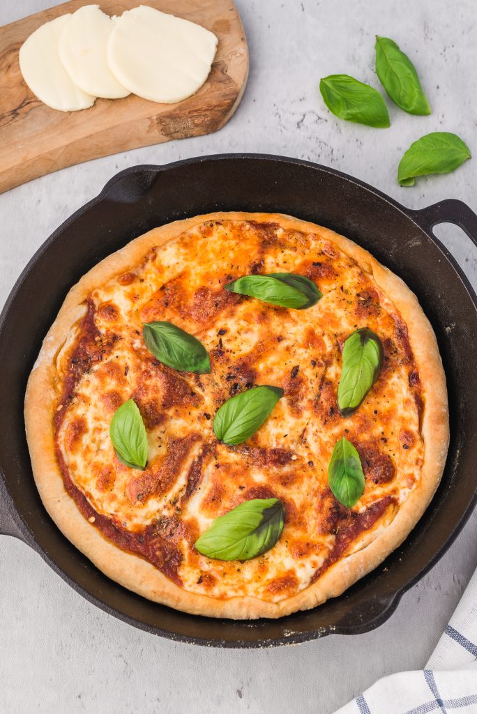 A whole cast iron pizza in the pan with some sliced mozzarella and a few basil leaves off to the side.