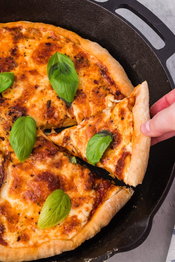 A hand lifts a slice of pizza from a cast iron pan.