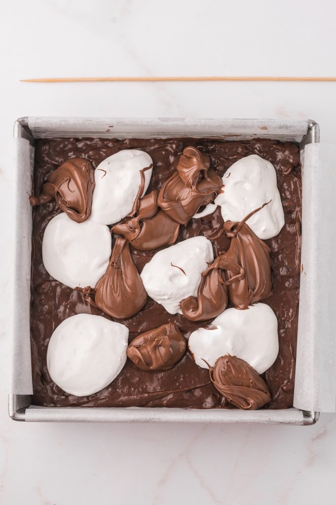 The mixed brownie batter topped with globs of nutella and marshmallow fluff in a parchment lined brownie tray.