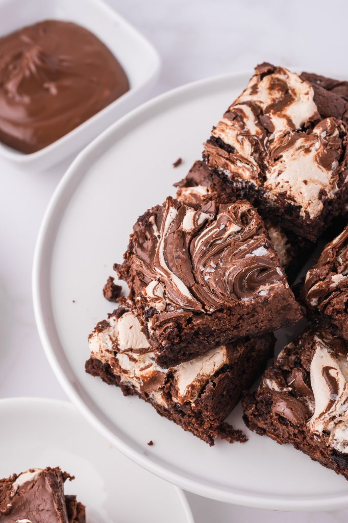 A pile of nutella fluff brownies on a white plate paper next to a bowl of nutella.