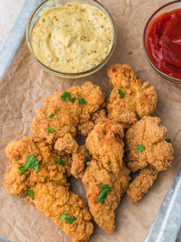 A tray of cooked Air Fryer Frozen Chicken Tenders with mustard and ketchup in two small glass bowls.