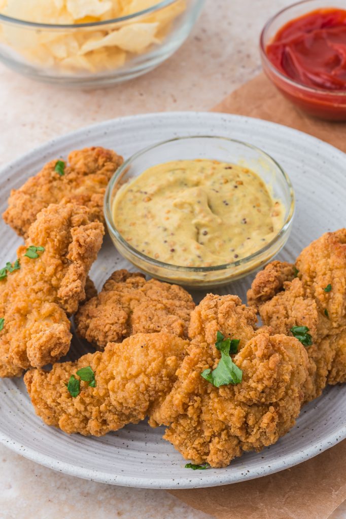 A plate of cooked Air Fryer Frozen Chicken Tenders next to a small glass bowl of mustard.