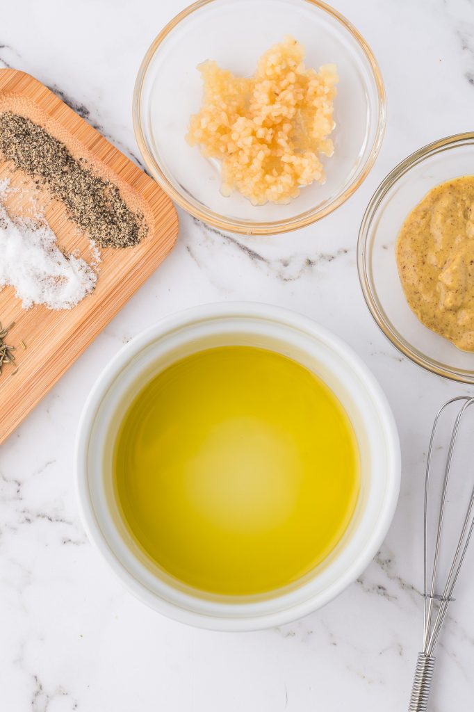 Oil, garlic, and dijon mustard in small glass bowls next to a plate of herbs.