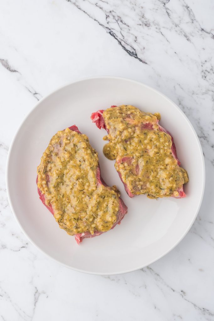 Two raw steaks topped with the dijon mixture on a white plate.