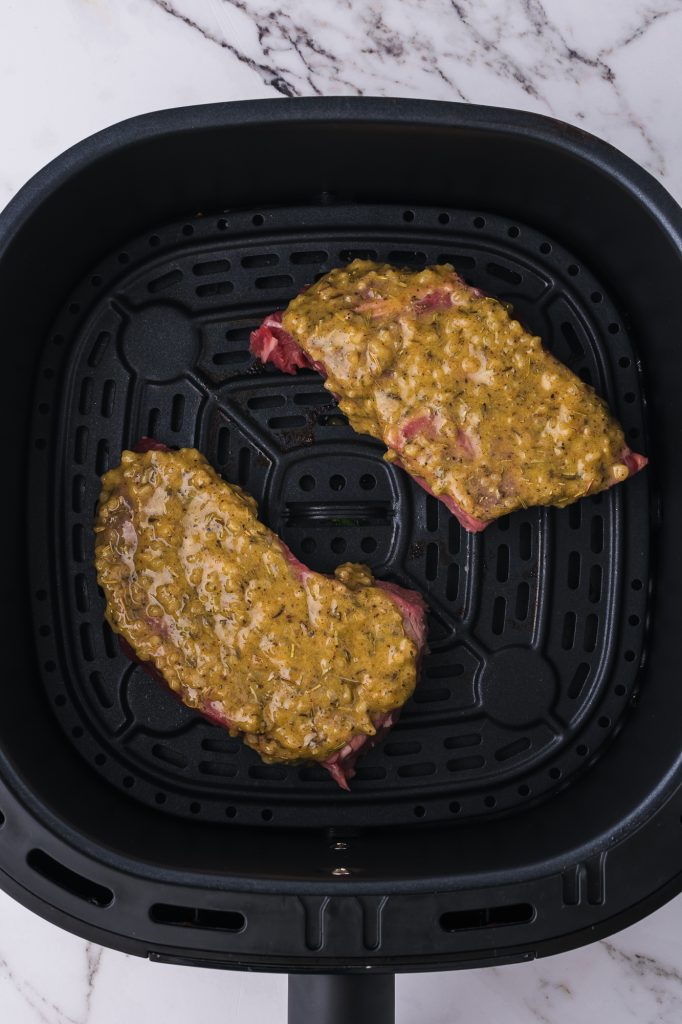 Two raw steaks topped with the dijon mixture in an air fryer basket.