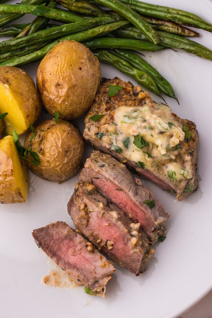 A sliced Air Fryer Steak with Garlic Butter on a white plate next to some boiled potatoes and roasted green beans.