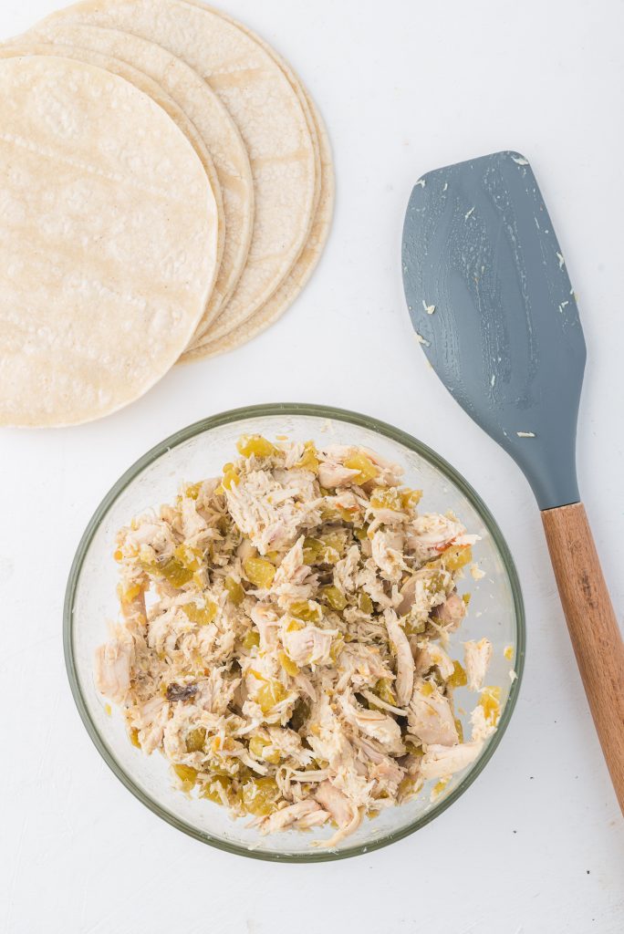 The chicken and chile mixture in a glass bowl next to a pile of tortillas and a spatula.