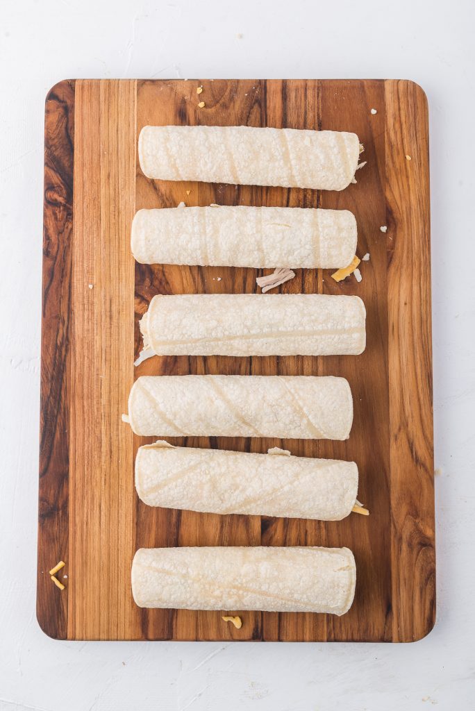 A vertical row of six Air Fryer Chicken and Cheese Taquitos on a wooden cutting board.