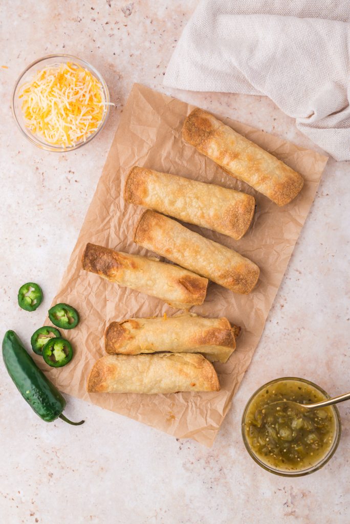 Six fully cooked Air Fryer Chicken and Cheese Taquitos on a piece of parchment paper with a bowl of shredded cheese, a bowl of salsa verde, and some jalapeno peppers next to it.