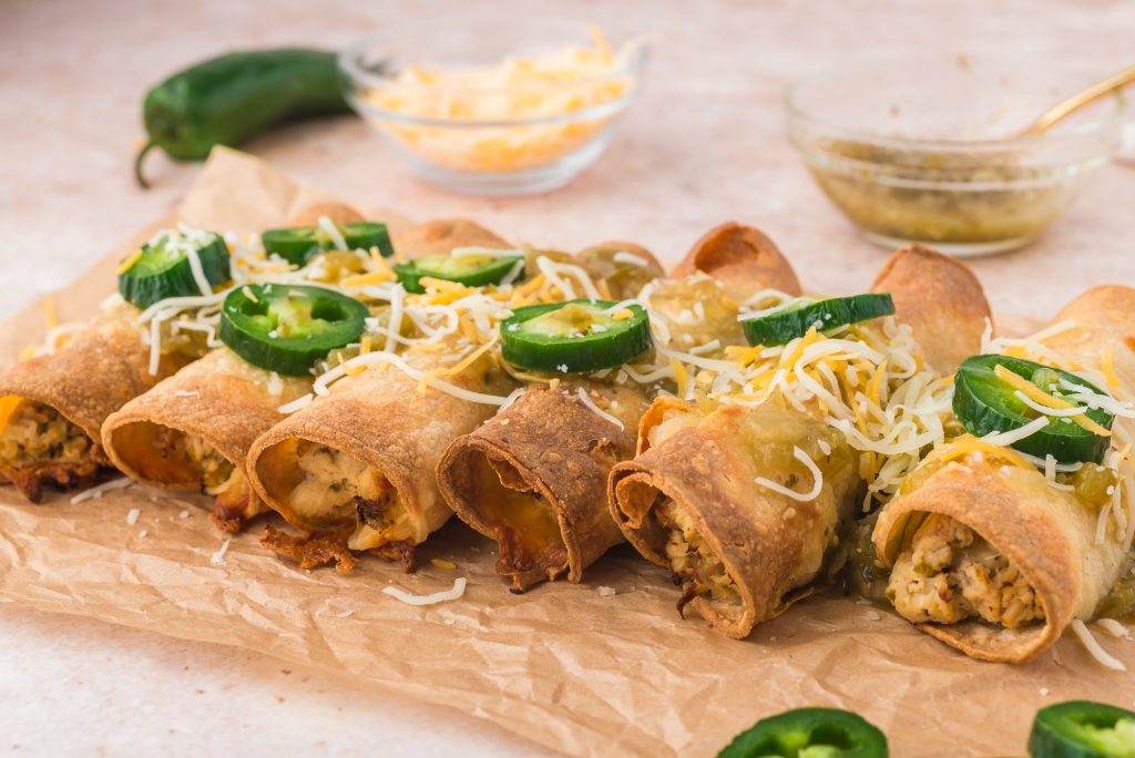 A row of Air Fryer Chicken and Cheese Taquitos topped with salsa verde, shredded cheese, and sliced jalapeno peppers.