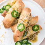 A plate of six Air Fryer Chicken and Cheese Taquitos topped with shredded cheese and sliced jalapeno peppers.