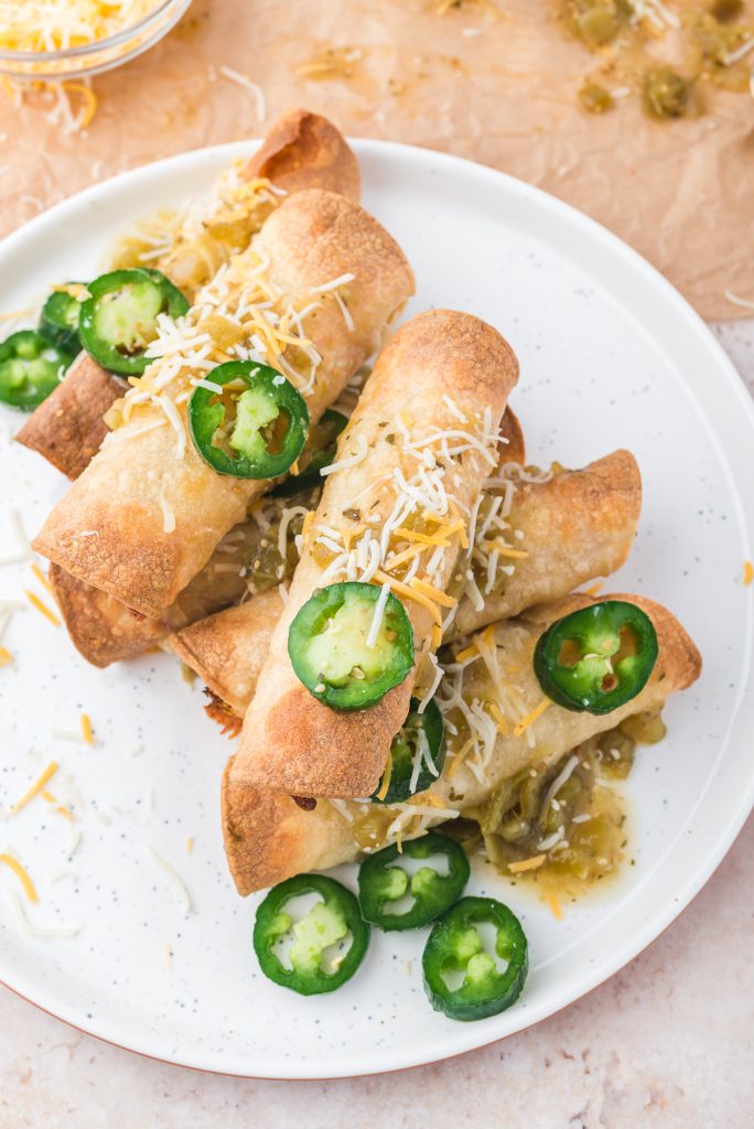 A plate of six Air Fryer Chicken and Cheese Taquitos topped with shredded cheese and sliced jalapeno peppers.