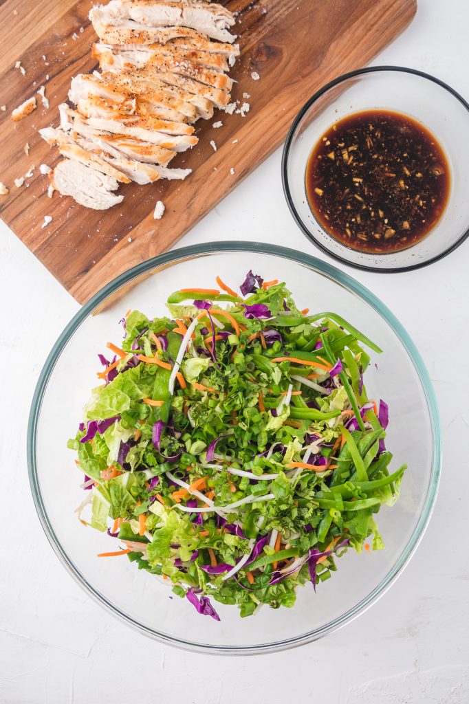 The combined salad in a glass bowl, next to the dressing in a glass bowl, next to the grilled chicken on a wooden cutting board.