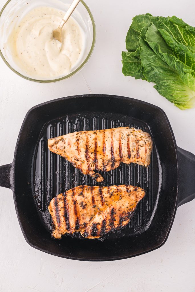 Two chicken breasts grilling on a skillet with char marks.