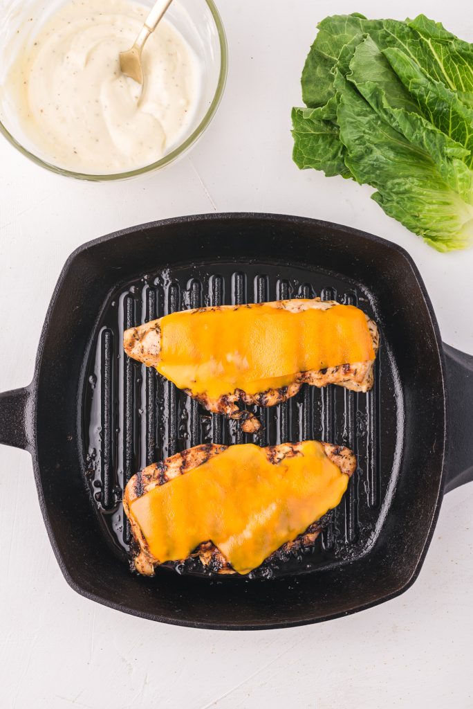 Two chicken breasts grilling on a skillet with char marks topped with melted cheddar cheese.