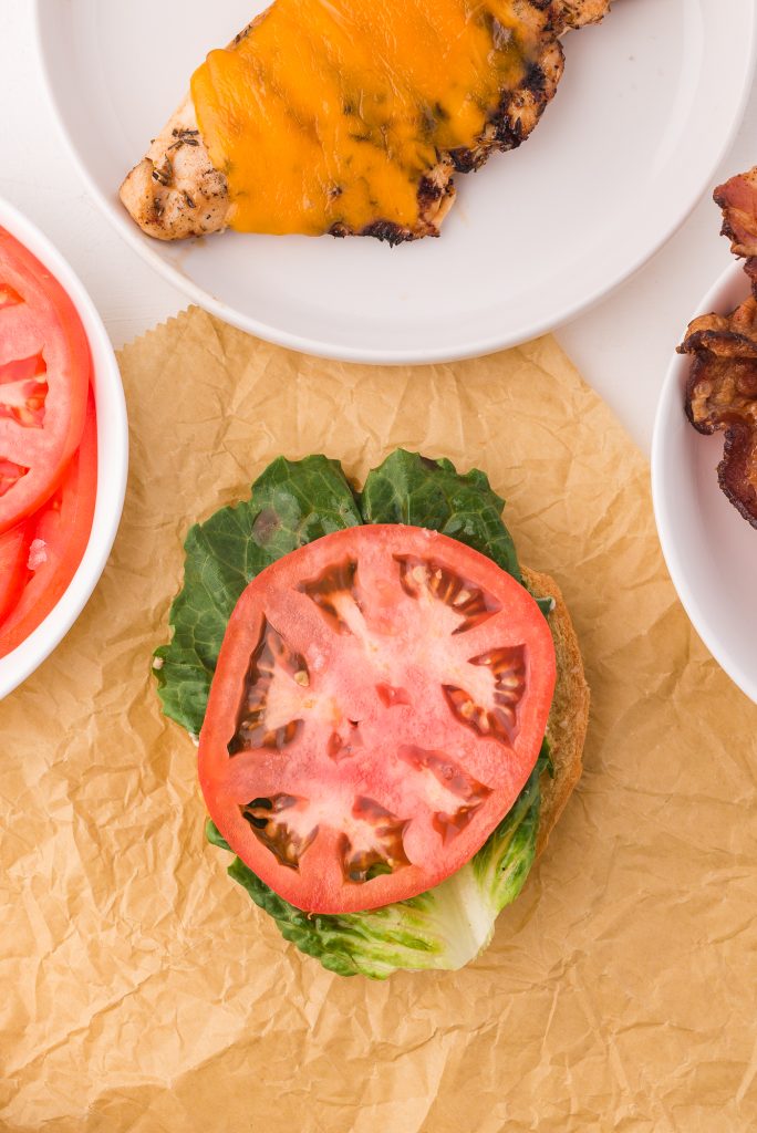 A split hamburger bun topped with garlic aioli, a piece of lettuce, then a slice of tomato.