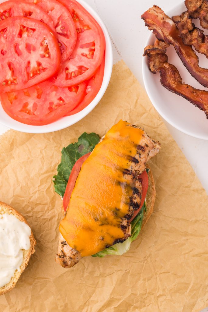 A split hamburger bun topped with garlic aioli, a piece of lettuce, a slice of tomato, then the grilled chicken with melted cheese on top.