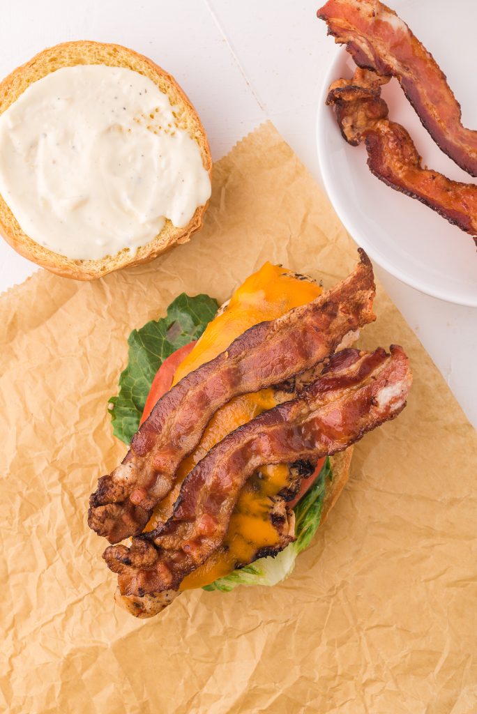 A split hamburger bun topped with garlic aioli, a piece of lettuce, a slice of tomato, the grilled chicken with melted cheese on top, then two strips of crisp bacon.