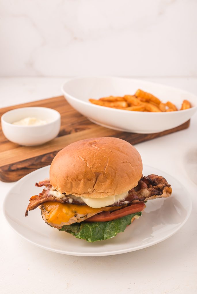 A grilled chicken sandwich with garlic aioli on a white plate with a wooden board with fries and more garlic aioli in the background.