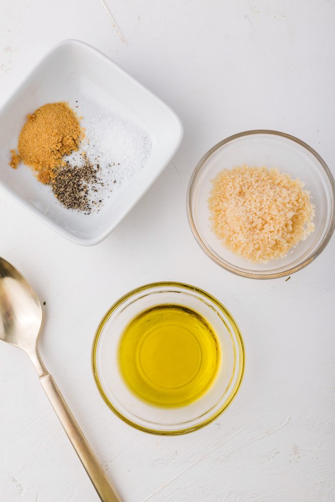 Three bowls containing spices, parmesan cheese, and oil, and a spoon rests next to them.