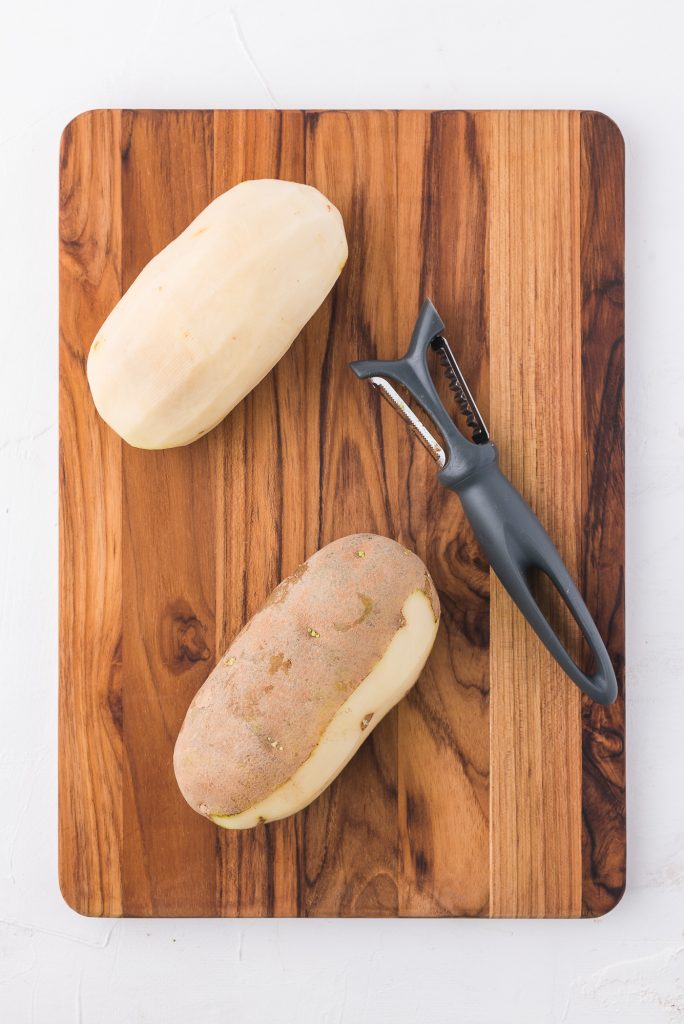 One fully peeled potoato and one half peeled potato sitting on a wooden cutting board with a vegetable peeler.