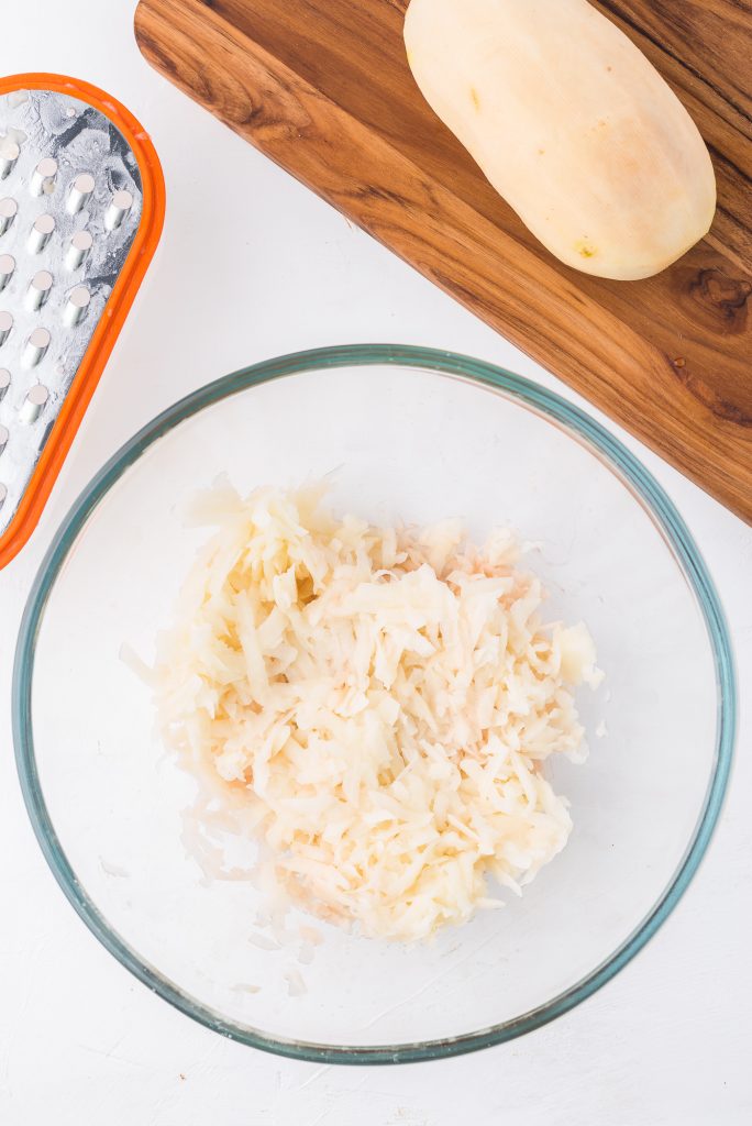 A glass bowl of grated potato sitting next to a grater that is mostly out of the picture.