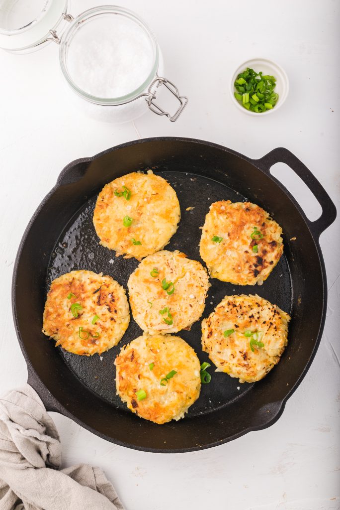 Six Cheesy Potato and Onion Breakfast Pancakes frying in a cast iron skillet.