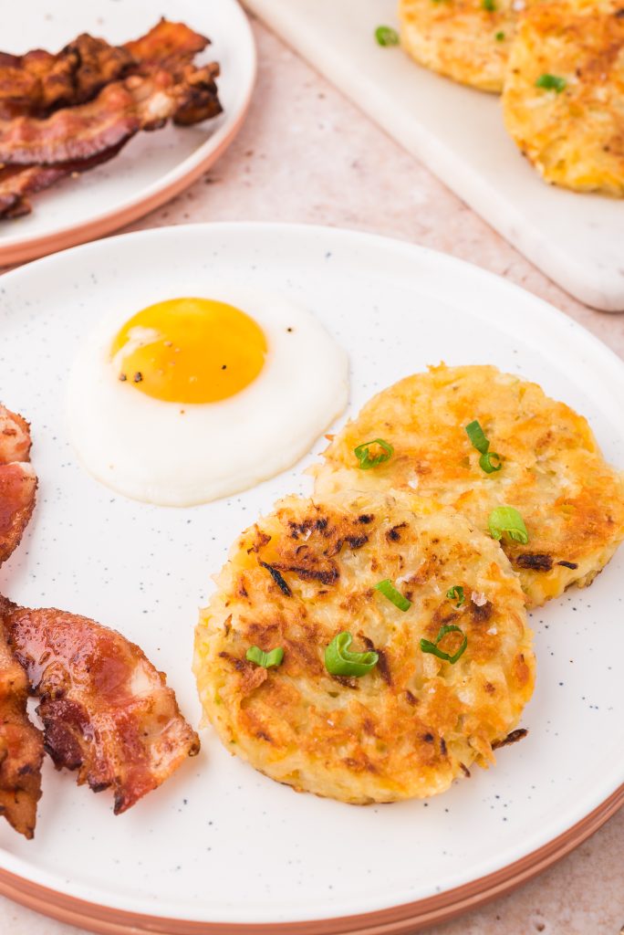 A white plate with two Cheesy Potato and Onion Breakfast Pancakes, a sunny side up egg, and two strips of crisp bacon.