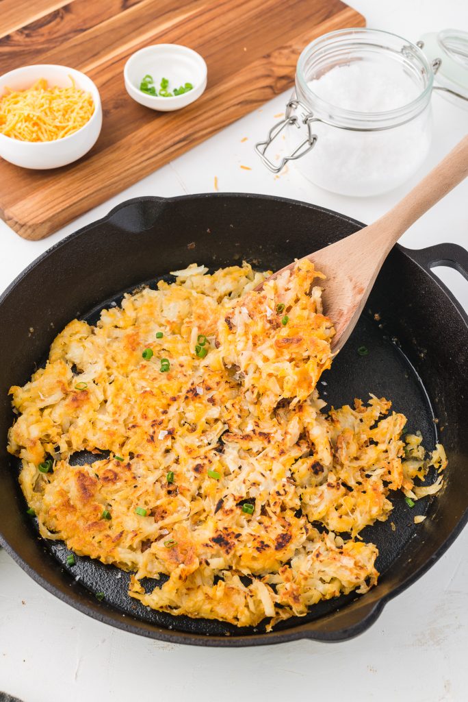 A wooden spoon about to serve the Quick Cheesy Homemade Hash Browns from a cast iron skillet.