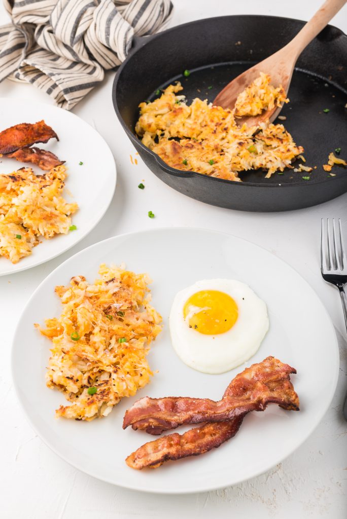 A serving of the Quick Cheesy Homemade Hash Browns on a white plate next to a sunny-side up egg and a couple strips of bacon. The skillet with the remainder hash browns is in the background.