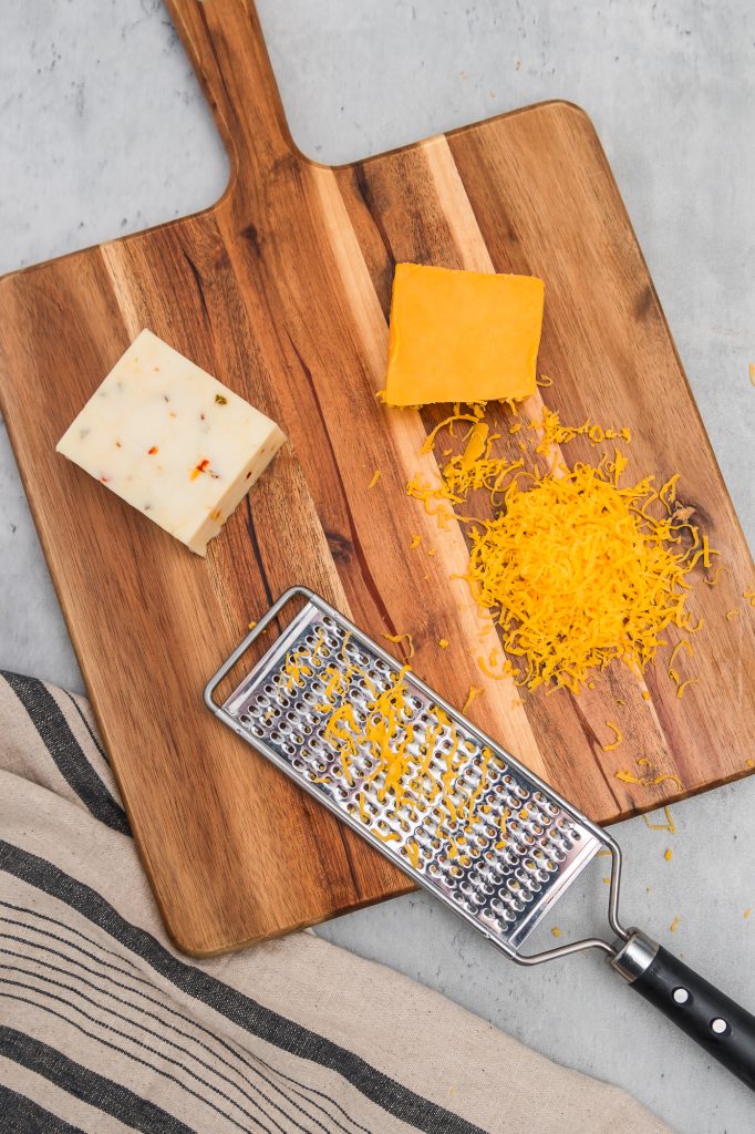 The cheeses being grated on a grater on a wooden cutting board.