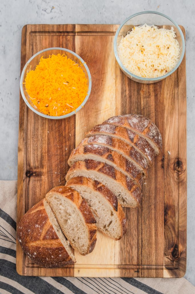 The shredded cheese and the sliced bread on a wooden cutting board.