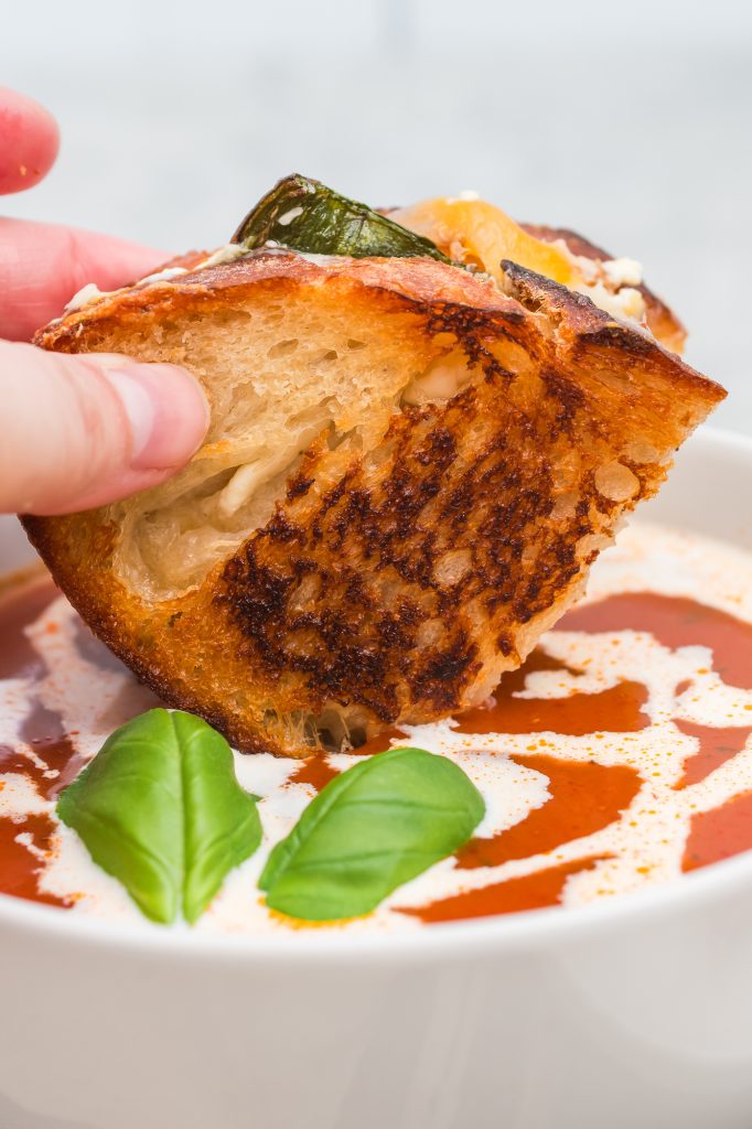 A closeup of a half of a Jalapeño Grilled Cheese Sandwich being dipped into a bowl of tomato soup.