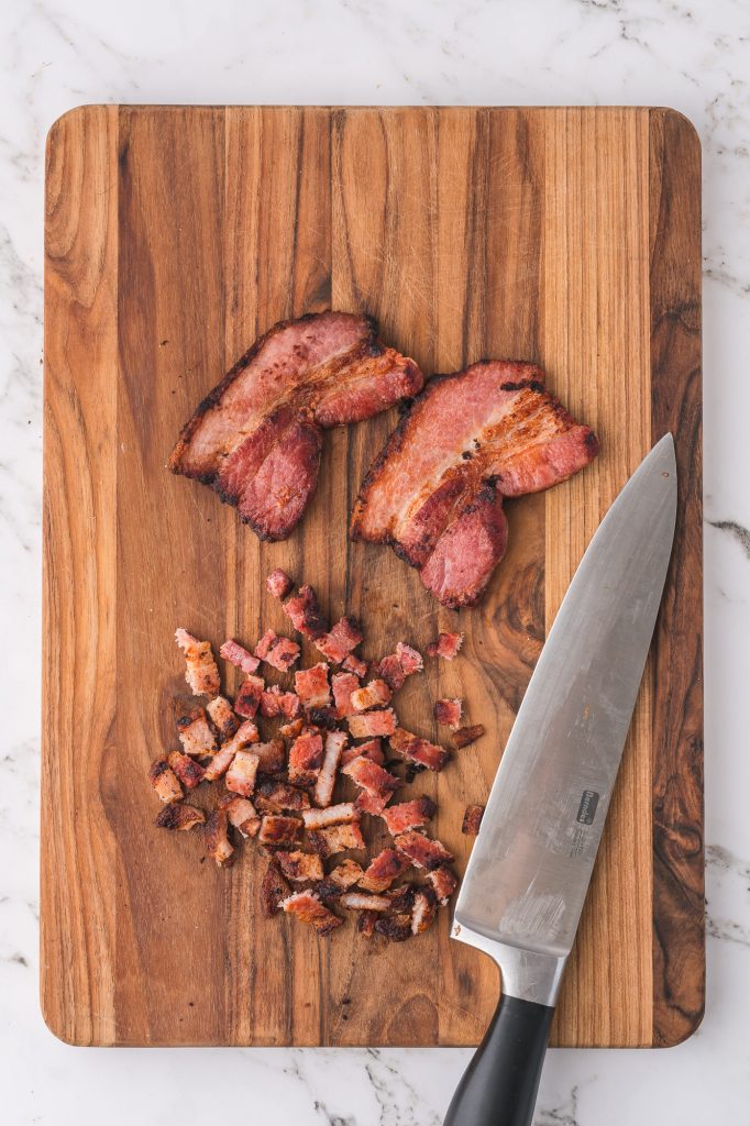 Cooked bacon on a wooden cutting board getting chopped into pieces.