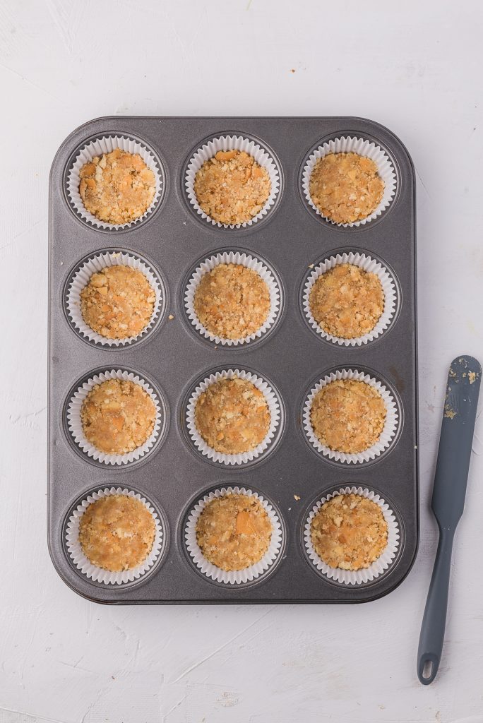 A muffin tin lined with paper cups and each one has crust mixture pressed into it.