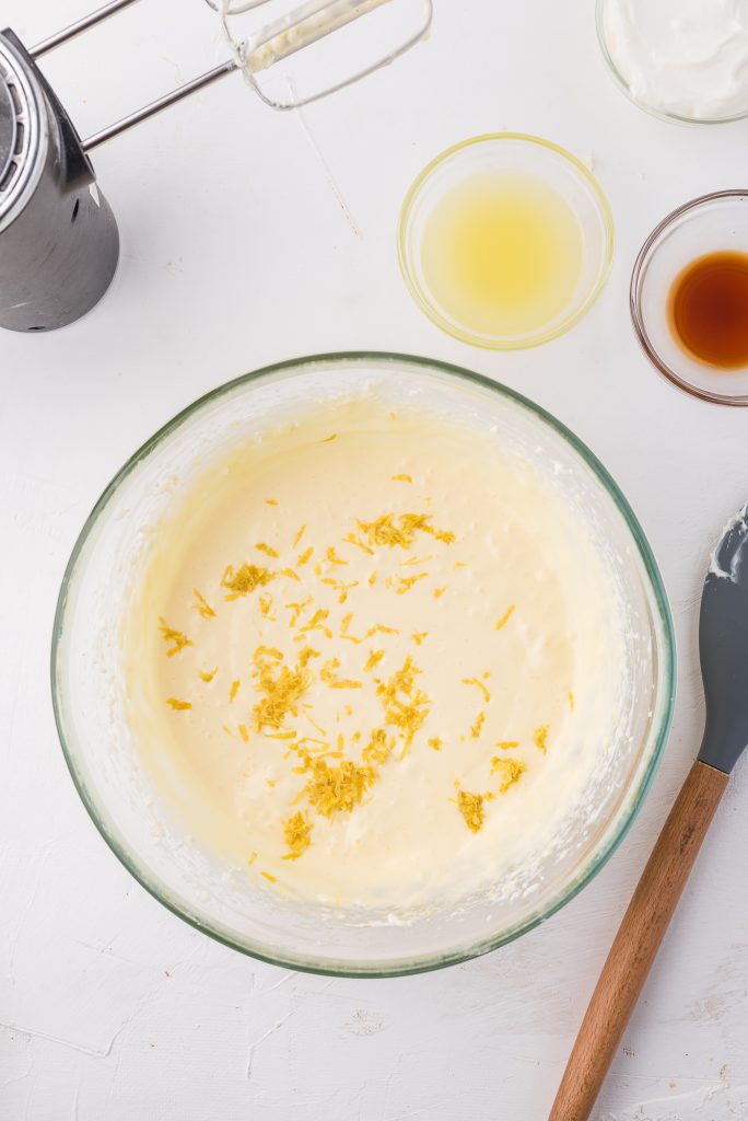 A cream cheese mixture with greek yogurt, lemon zest, lemon juice, and vanilla extract about to be stirred in until fully incorporated in a glass bowl.