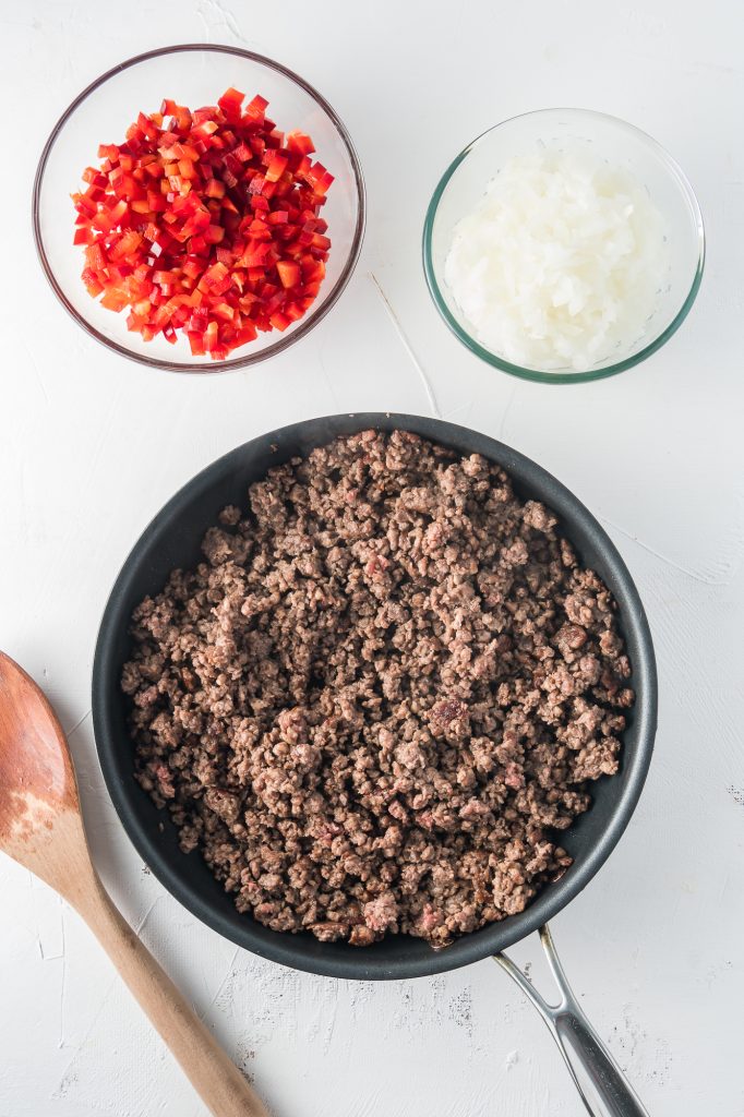 A pan of cooked ground beef with bowls of chopped onion and red pepper to the side of it.