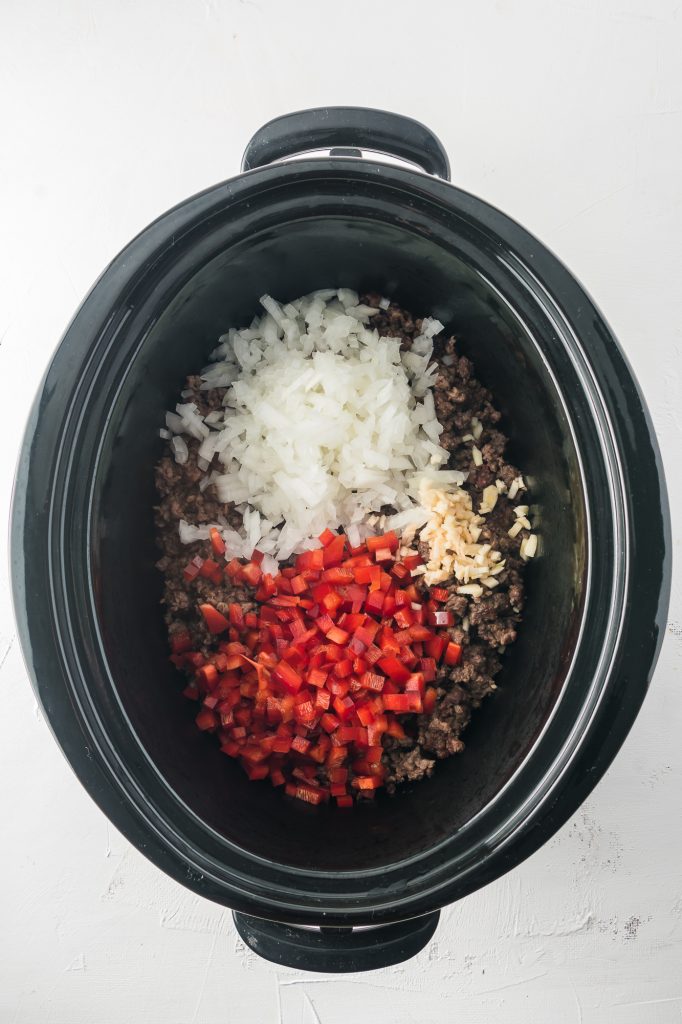 Cooked ground beef topped with chopped onion and red pepper at the bottom of a slow cooker.