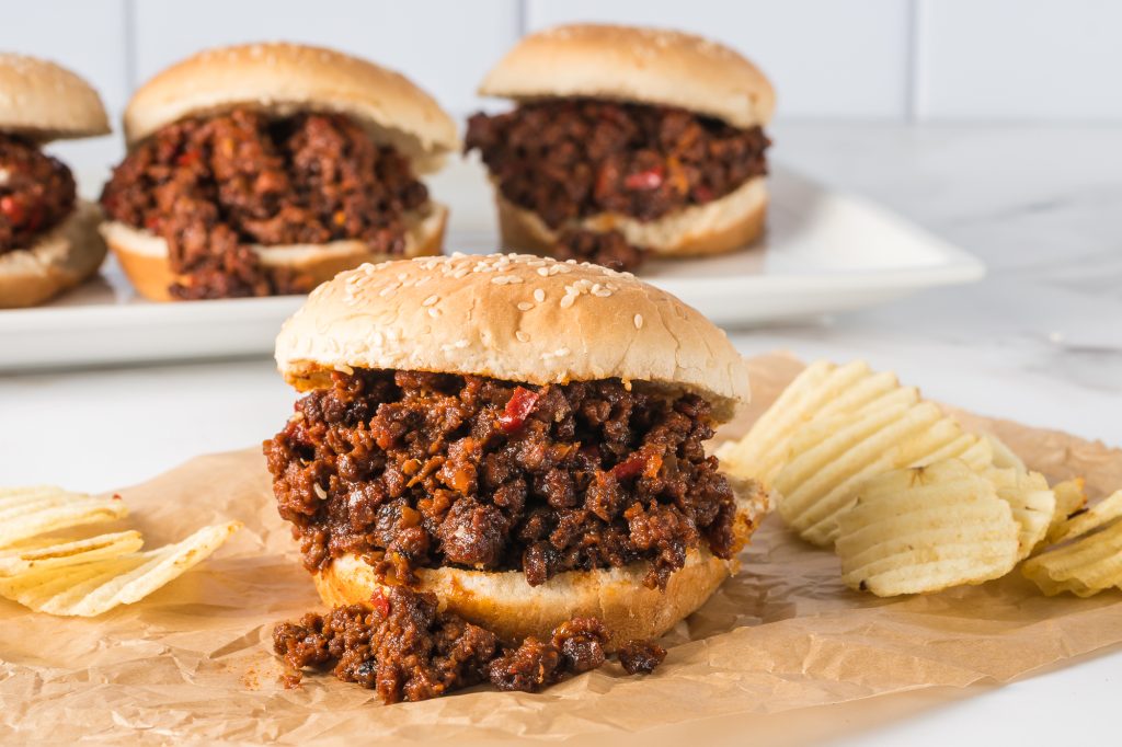 A completed sloppy joe on a piece of parchment paper next to a pile of potato chips. The other three completed sloppy joes are in the background on a white plate.