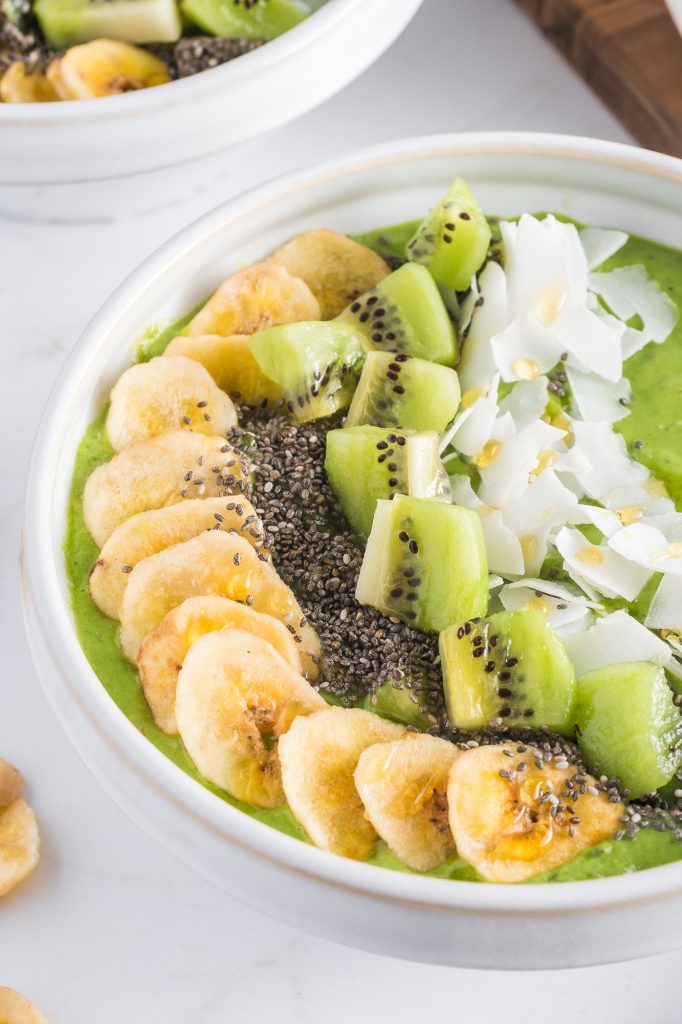 A close up of white bowl full of the Tropical Green Smoothie Bowl base topped with bananas, kiwi, chia seeds, coconut flakes.