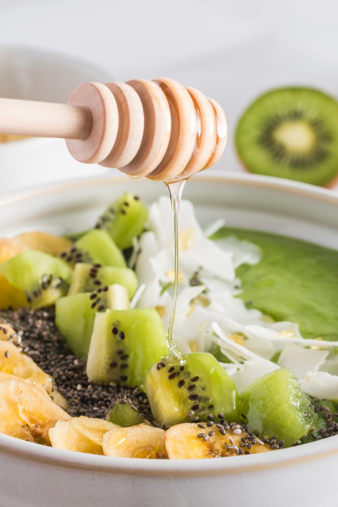 A close up of white bowl full of the Tropical Green Smoothie Bowl base topped with bananas, kiwi, chia seeds, coconut flakes being drizzled with honey.